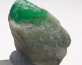 May include: A rough-cut emerald gemstone with a vibrant green hue on the top surface. The stone has a textured, irregular surface with a mix of green and gray tones. The gemstone is mounted on a small, gray base.