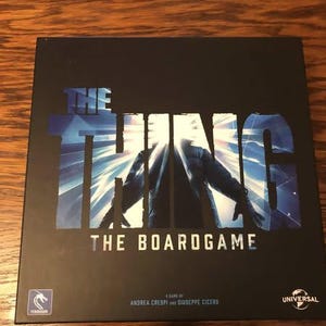 May include: The Thing board game box cover features a black background with a blue and white graphic of the creature from the movie. The text "The Thing The Boardgame" is printed in white on the box. The game is by Andrea Crespi and Giuseppe Cicero.