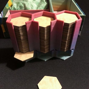 May include: A close-up of a hexagonal game tile organizer with three compartments. The organizer is made of a pink and purple plastic and is filled with hexagonal game tiles. The tiles are stacked neatly in each compartment. The organizer is sitting on a black surface.