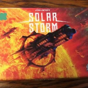 May include: A box for the board game "Solar Storm" by Aiden Lowther's. The box features a fiery orange and red background with a spaceship flying through a solar flare. The game logo is in white and black.