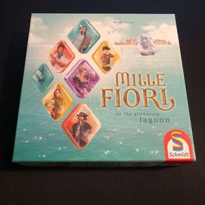 May include: A blue and green board game box with the title "Mille Fiori: In the glittering lagoon" and a colourful illustration of people and a boat on the water.