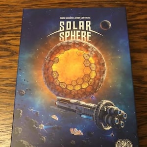 May include: A blue box with a space-themed design. The box features a large, orange, honeycomb-like planet with a spaceship flying in front of it. The text "Solar Sphere" is printed in white on the box. The box is from "Dkanua Games".