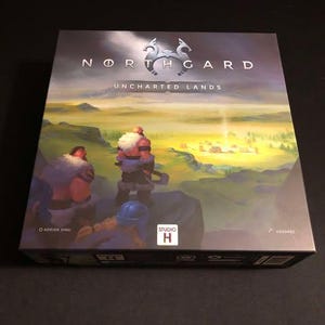 Puede incluir: Una caja de juego de mesa con un fondo azul oscuro y un logotipo plateado de dos animales estilizados. El texto "Northgard Uncharted Lands" está impreso en blanco en la caja. La caja tiene una ilustración azul claro y blanco de un paisaje con dos figuras en primer plano.