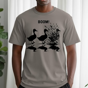 May include: A gray t-shirt with a black graphic of three ducks, one exploding with the word "BOOM!" above. The design includes stylized water splashes and duck silhouettes. The shirt is a casual wear item.