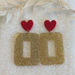 Peut inclure: Boucles d'oreilles rectangulaires dorées à paillettes avec un cœur rouge en haut. Les boucles d'oreilles ont une découpe rectangulaire au centre. Les boucles d'oreilles sont suspendues à un crochet doré. Le fond est blanc.