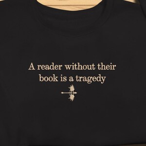 Op de afbeelding: Zwart sweatshirt met de quote "A reader without their book is a tragedy" in een schreeflettertype. Onder de tekst staat een kleine afbeelding van een pijl en boog. Het sweatshirt ligt op een houten ondergrond.