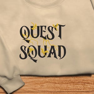 May include: A beige sweatshirt with the text "QUEST SQUAD" in black lettering. The text is surrounded by small yellow lightning bolts and birds.