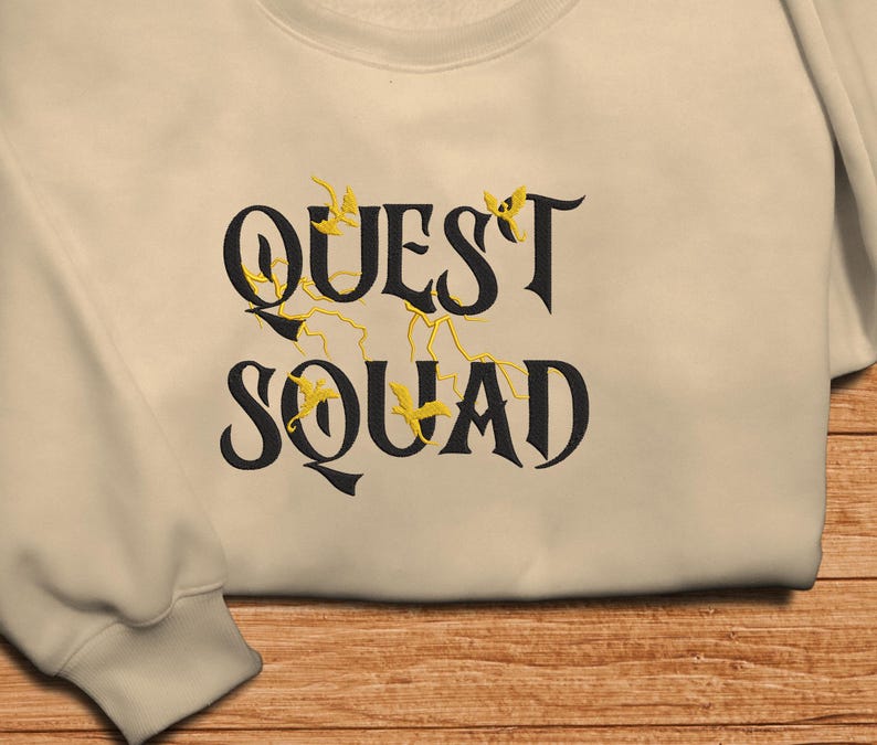 May include: A beige sweatshirt with the text "QUEST SQUAD" in black lettering. The text is surrounded by small yellow lightning bolts and birds.