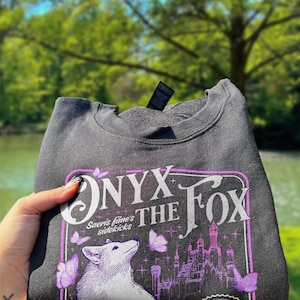 Może przedstawiać: Ciemnoszara bluza z fioletowo-białą grafiką. Grafika zawiera tekst "ONYX THE FOX" i "Saeris fane's sidekicks", białego lisa, motyle i zamek. Bluza jest złożona.