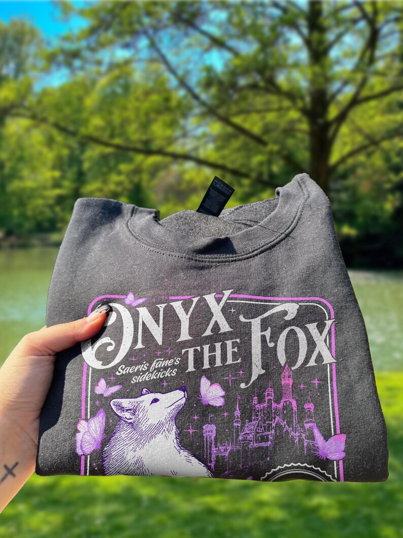 Może przedstawiać: Ciemnoszara bluza z fioletowo-białą grafiką. Grafika zawiera tekst "ONYX THE FOX" i "Saeris fane's sidekicks", białego lisa, motyle i zamek. Bluza jest złożona.