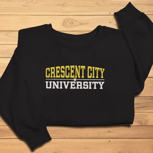 May include: A black sweatshirt with the text "CRESCENT CITY UNIVERSITY" in yellow and white block letters. The sweatshirt is made of a soft material and is displayed on a wooden surface.