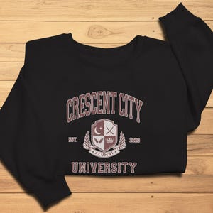 May include: Black sweatshirt with the text "CRESCENT CITY UNIVERSITY ALUMNI" in burgundy. The design includes a crest with a crescent moon, crossed swords, and a crown. The year "2020" is also displayed.
