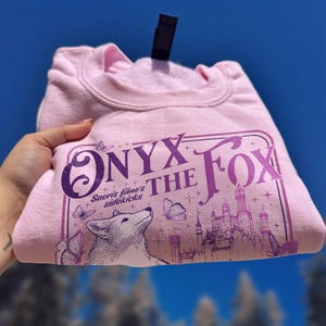 Może przedstawiać: Jasnor&oacute;żowa bluza z napisem "ONYX THE FOX" w kolorze fioletowym. Projekt zawiera lisa, motyle i zamek. Złożona bluza jest prezentowana na tle błękitnego nieba, z widoczną czarną metką.