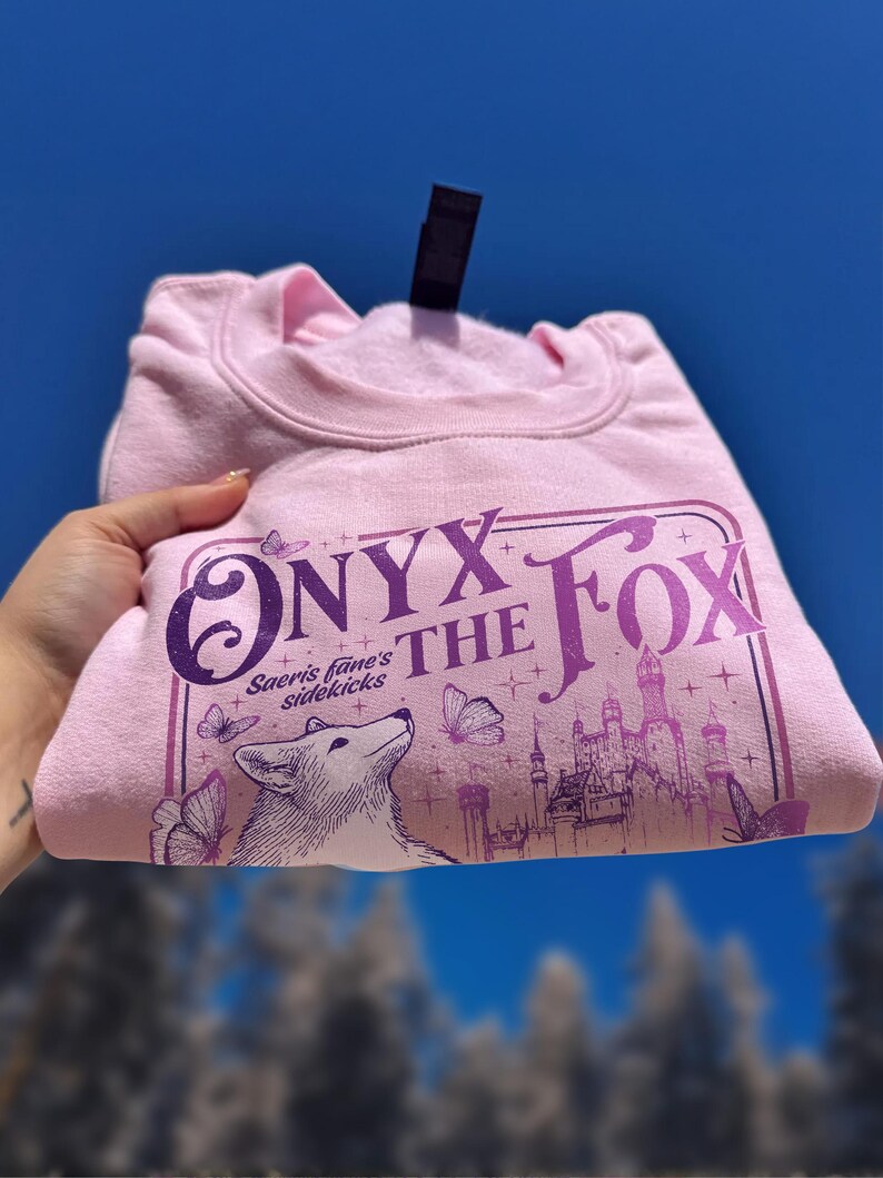 Może przedstawiać: Jasnor&oacute;żowa bluza z napisem "ONYX THE FOX" w kolorze fioletowym. Projekt zawiera lisa, motyle i zamek. Złożona bluza jest prezentowana na tle błękitnego nieba, z widoczną czarną metką.