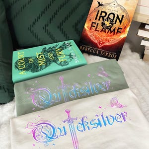 Pu&ograve; includere: Due magliette con la scritta "Quicksilver" in un font fantasy, una spada e grafiche di farfalle. Le magliette sono verde chiaro e bianco sporco. Sono visibili due libri, "A Court of Mist and Fury" e "Iron Flame" di Rebecca Yarros.