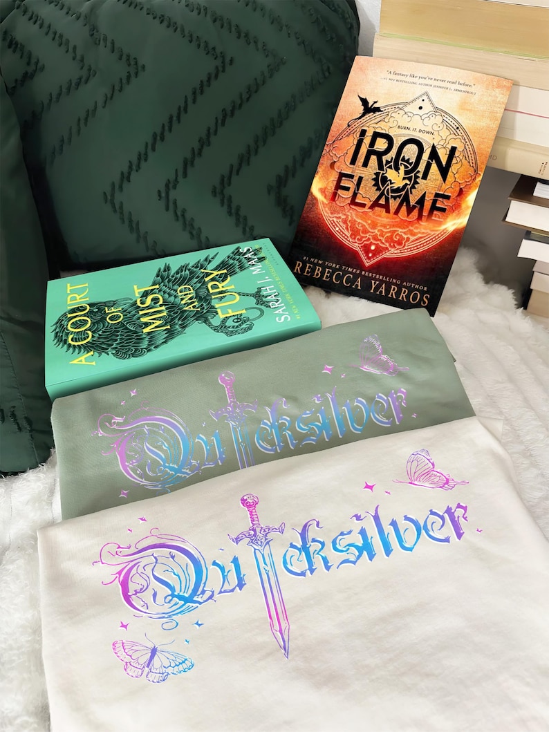 Pu&ograve; includere: Due magliette con la scritta "Quicksilver" in un font fantasy, una spada e grafiche di farfalle. Le magliette sono verde chiaro e bianco sporco. Sono visibili due libri, "A Court of Mist and Fury" e "Iron Flame" di Rebecca Yarros.