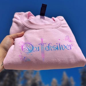 Pu&ograve; includere: Una felpa rosa chiaro con la scritta "Quicksilver" in un gradiente blu e viola, insieme a grafiche di spada e farfalla. La felpa &egrave; piegata e tenuta contro uno sfondo di cielo blu.