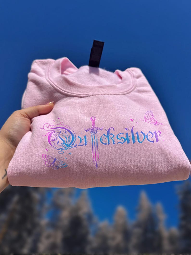 Pu&ograve; includere: Una felpa rosa chiaro con la scritta "Quicksilver" in un gradiente blu e viola, insieme a grafiche di spada e farfalla. La felpa &egrave; piegata e tenuta contro uno sfondo di cielo blu.