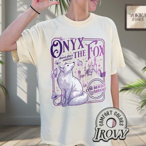 Może przedstawiać: Kremowa koszulka z fioletową grafiką lisa, napisem "ONYX THE FOX" i "The Bravest Warrior". Projekt zawiera motyle i ilustrację zamku.
