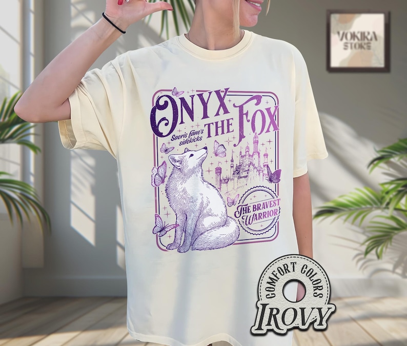 Może przedstawiać: Kremowa koszulka z fioletową grafiką lisa, napisem "ONYX THE FOX" i "The Bravest Warrior". Projekt zawiera motyle i ilustrację zamku.