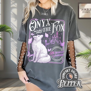 Może przedstawiać: Ciemnoszara koszulka z ilustracją białego lisa i napisem "ONYX THE FOX" w fioletowej ramce. Projekt zawiera motyle i zamek. Koszulka ma wygląd vintage z napisem "COMFORT COLORS PEPPER" na dole.