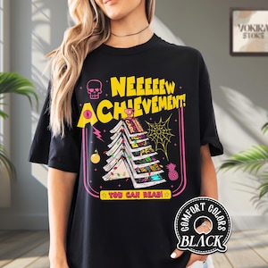 May include: Black t-shirt with a graphic design featuring the text "NEEEEEEW ACHIEVEMENT!" in yellow, a skull, and a Christmas tree made of books. The design includes a spiderweb, a pineapple, and the words "YOU CAN READ!"