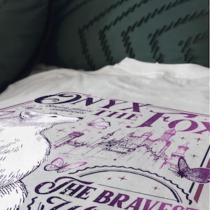 Może przedstawiać: Biała koszulka z fioletowym nadrukiem graficznym. Projekt zawiera tekst "Onyx the Fox" i "The Bravest Warrior", ilustrację lisa, motyle i zamek. Koszulka wykonana jest z miękkiego, wygodnego materiału.