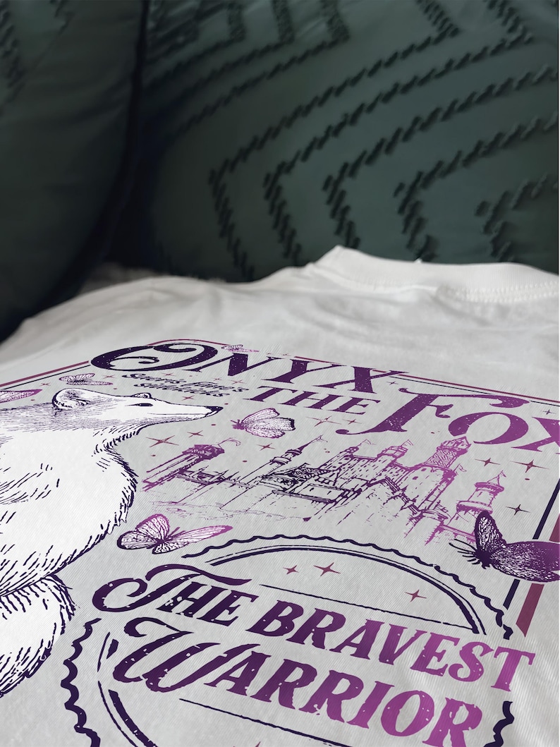 Może przedstawiać: Biała koszulka z fioletowym nadrukiem graficznym. Projekt zawiera tekst "Onyx the Fox" i "The Bravest Warrior", ilustrację lisa, motyle i zamek. Koszulka wykonana jest z miękkiego, wygodnego materiału.