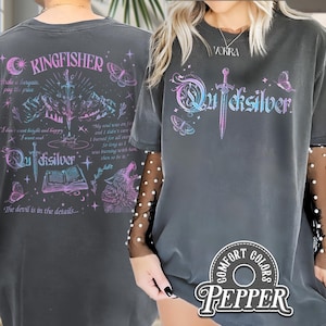 Pu&ograve; includere: T-shirt Comfort Colors grigio scuro con un design grafico. Il design presenta la parola "Quicksilver" in un font stilizzato, una spada, farfalle e la parola "Kingfisher". La maglietta ha un aspetto vintage e usurato.