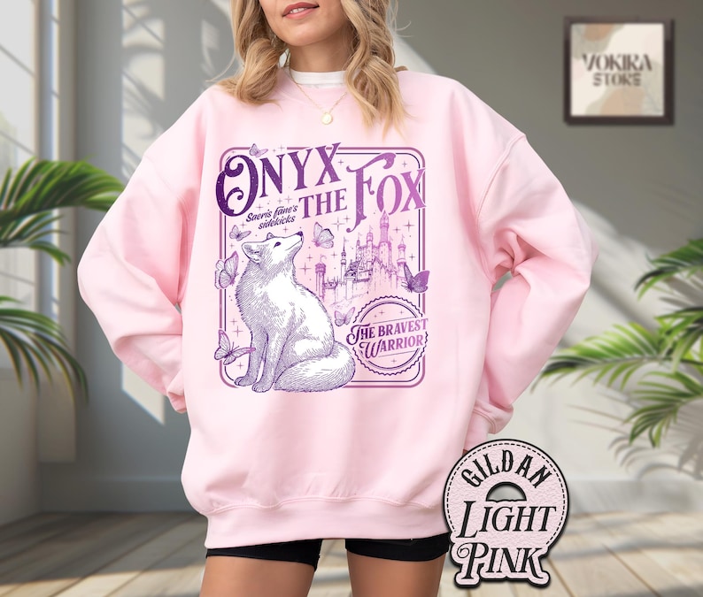 Może przedstawiać: Jasnor&oacute;żowa bluza z fioletową grafiką lisa, napisem "Onyx the Fox" i "The Bravest Warrior". Projekt zawiera motyle i zamek. Bluza to Gildan Light Pink.