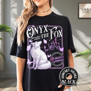 Może przedstawiać: Czarny t-shirt z ilustracją białego lisa i napisem "ONYX THE FOX" w fioletowej ramce. Projekt zawiera motyle i zamek, z tekstem "THE BRAVEST WARRIOR". Koszulka jest oznaczona jako "COMFORT COLORS BLACK".