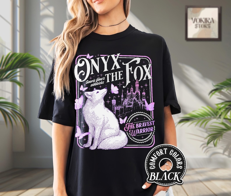Może przedstawiać: Czarny t-shirt z ilustracją białego lisa i napisem "ONYX THE FOX" w fioletowej ramce. Projekt zawiera motyle i zamek, z tekstem "THE BRAVEST WARRIOR". Koszulka jest oznaczona jako "COMFORT COLORS BLACK".