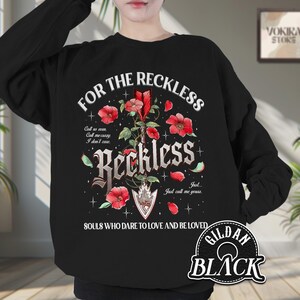 Può includere: Felpa nera con la scritta "FOR THE RECKLESS" e "Reckless" in un font gotico. Il design include fiori rossi, foglie e le parole "SOULS WHO DARE TO LOVE AND BE LOVED".