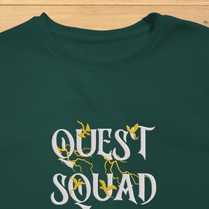 May include: A dark green sweatshirt with the text "QUEST SQUAD" in white lettering with yellow accents. The text is in a stylized font.