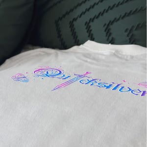 Pu&ograve; includere: T-shirt bianca con un design colorato con la scritta "Quicksilver" in un font stilizzato, una spada e illustrazioni di farfalle. Il design utilizza sfumature di blu, viola e rosa. La maglietta &egrave; realizzata in morbido tessuto bianco.