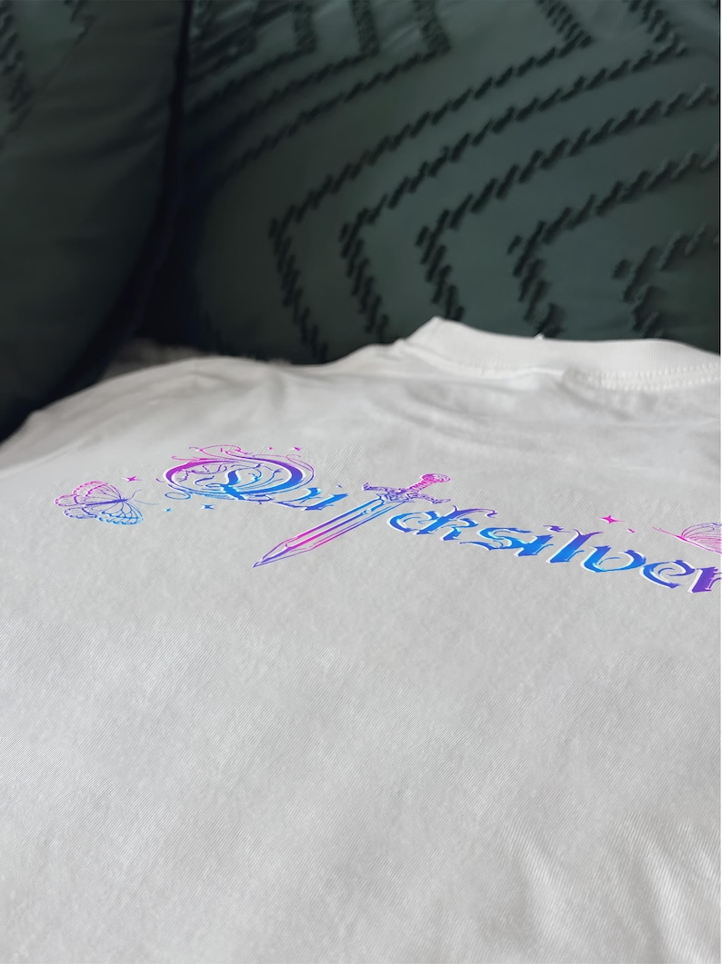 Pu&ograve; includere: T-shirt bianca con un design colorato con la scritta "Quicksilver" in un font stilizzato, una spada e illustrazioni di farfalle. Il design utilizza sfumature di blu, viola e rosa. La maglietta &egrave; realizzata in morbido tessuto bianco.