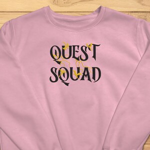 May include: Pink sweatshirt with the text "QUEST SQUAD" in black lettering with gold accents.