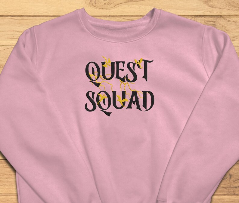 May include: Pink sweatshirt with the text "QUEST SQUAD" in black lettering with gold accents.