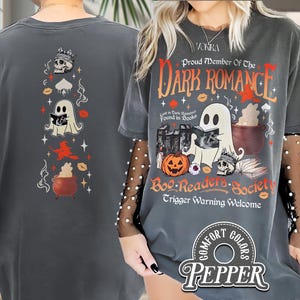 Puede incluir: Camiseta gris oscuro Comfort Colors con un diseño de Halloween. La parte delantera presenta un fantasma leyendo un libro, una calabaza y el texto "Proud Member of the Dark Romance Boo Readers Society". La parte trasera tiene una calavera, un fantasma, una bruja y un caldero.