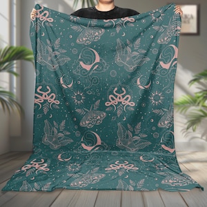 Peut inclure: Une couverture de couleur sarcelle avec un motif répété de papillons de nuit, de lunes et de motifs célestes en rose pâle. La couverture est tenue, mettant en valeur le design complet et la texture du tissu.