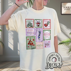 May include: Cream-colored t-shirt featuring a collage of postage stamp designs. The stamps showcase various illustrations, including a castle, a heart, flowers, and crowns, with phrases like "Vicious Little Thing" and "Powerless". The shirt has a relaxed fit.