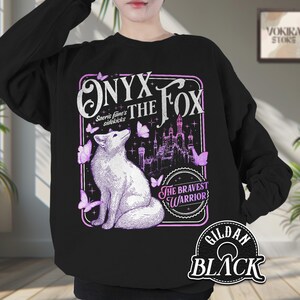 Może przedstawiać: Czarna bluza z ilustracją białego lisa, napisem "Onyx the Fox" i "The Bravest Warrior". Projekt zawiera zamek i fioletowe motyle. Bluza to Gildan Black.