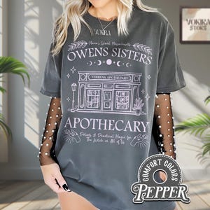 Może przedstawiać: Ciemnoszara koszulka z nadrukiem graficznym z napisem "Owens Sisters Apothecary" oraz słowami "Maria's Island, Massachusetts". Projekt zawiera ilustracje fasady sklepu, gwiazd i księżyców. Koszulka ma etykietę Comfort Colors.