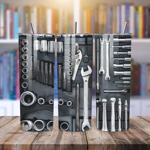 May include: Three stainless steel tumblers with a design of various tools, including wrenches, screwdrivers, and sockets. The tumblers are on a wooden surface.