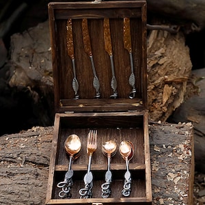 Puede incluir: Un juego de cubiertos en una caja de madera. El juego incluye cuatro cuchillos con hojas doradas texturizadas y mangos en forma de hoja, junto con cucharas y un tenedor. La cubertería tiene un acabado cobrizo y detalles decorativos de hojas.