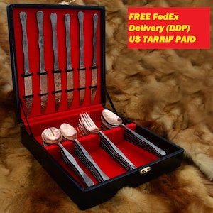 May include: A flatware set featuring six knives with hammered copper-toned blades and six matching spoons and forks, all housed in a black presentation box with a vibrant red interior lining. The set includes a gold-toned metal latch closure.