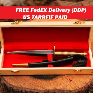 May include: A dagger with a silver blade and gold handle, presented in a wooden box with a red velvet interior. The dagger is accompanied by a black sheath with gold accents. The box is closed with gold clasps.
