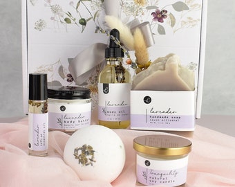 Könnte beinhalten: Ein Geschenkset zum Thema Lavendel mit einer weißen Box, einem Lavendel-Körperöl, einer Lavendel-Körperbutter, einer handgefertigten Lavendel-Seife, einer Lavendel-Badebombe und einer Lavendel-Sojawachskerze. Die Box ist mit einem Blumenmuster und einem grauen Band verziert.