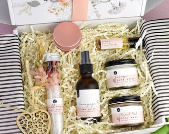 Könnte beinhalten: Eine Geschenkbox gefüllt mit verschiedenen Produkten zur Selbstpflege, darunter ein Badesalz, eine Körperbutter, eine Tonerdemaske, ein Leinenspray und ein ätherisches Öl. Die Box ist mit zerkleinertem Papier ausgekleidet und mit einem rosa Band verziert.
