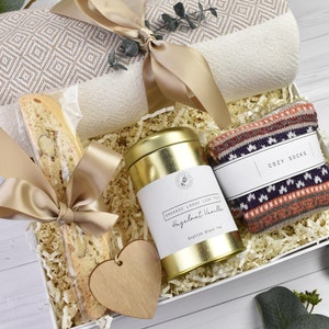 May include: A white gift box filled with a cosy throw blanket, a tin of Hazelnut Vanilla tea, a biscotti, a pair of cosy socks, and a wooden heart. The box is filled with shredded paper.